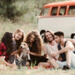 A group of young friends with a dog sitting on grass on a roadtrip through countryside.